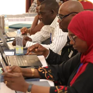 Early-career researchers seated at a conference table work on laptops during a training workshop, focusing on writing and peer review, with notebooks and water bottles in front of them.