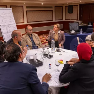 Participants in a roundtable workshop discuss ideas as a facilitator speaks, with notes on a flip chart and workshop materials spread across the table.