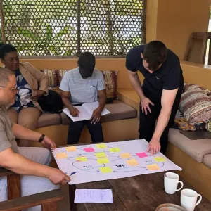 Four people sit and stand around a table during a small group workshop, reviewing a large paper diagram covered with sticky notes. The discussion appears collaborative and informal, taking place in a shaded outdoor or semi-open setting, with notebooks, cups, and writing materials visible.