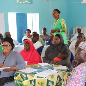 Men and women seated around tables facing left, listening. One woman, center rear, standing / speaking.