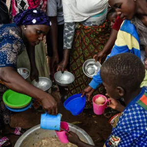 Woman, left, leans over metal pot of stew, lower center, pours stew from a cup into a cup held by a boy. Children, women surround them, some holding bowls.