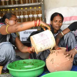 Three women seated on floor. One, left, pours bucket into bowl held by woman on right.