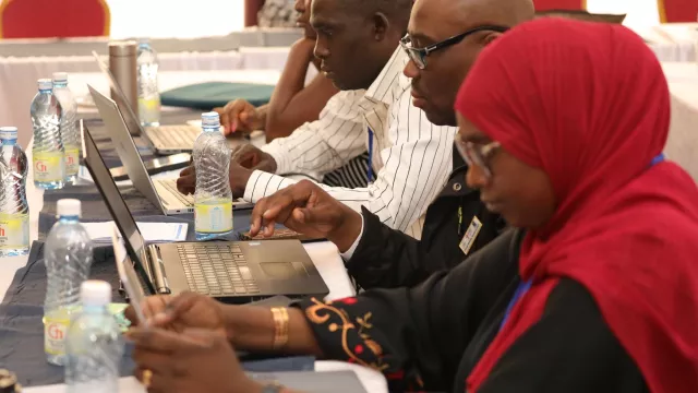 Early-career researchers seated at a conference table work on laptops during a training workshop, focusing on writing and peer review, with notebooks and water bottles in front of them.