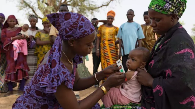 Woman, kneeling, left, measures baby's arm circumference. Baby is in arms of mother, right. Men and women stand in background.