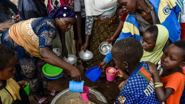 Woman, left, leans over metal pot of stew, lower center, pours stew from a cup into a cup held by a boy. Children, women surround them, some holding bowls.